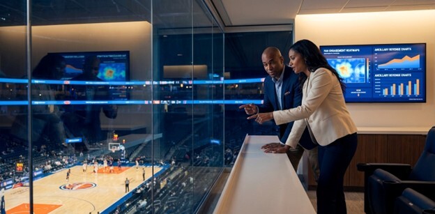 A female venue executive stands in a modern office overlooking a professional stadium at dusk. She is leaning over a desk covered in plans and financial charts, representing strategic planning and budget management. A large screen in the background shows data visualizations, underscoring the theme of using technology and data to solve complex operational challenges in the live events industry.