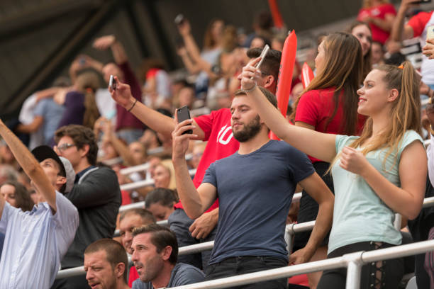 Fans in stadium stands cheering with smartphones raised, engaging with content on the jumbotron.